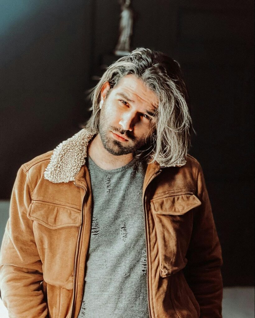 Stylish man with long hair wearing a brown jacket and grey shirt, exuding a serious expression.